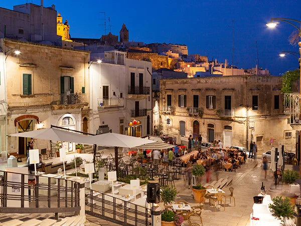 Abendliche Szene auf einer belebten Piazza in der malerischen apulischen Stadt Ostuni, bekannt als die 'Weiße Stadt', mit Gästen, die das Nachtleben bei einem Glas Primitivo-Wein genießen, während historische, weiß getünchte Gebäude im Hintergrund unter dem warmen Licht der Straßenlaternen und des blauen Abendhimmels leuchten.