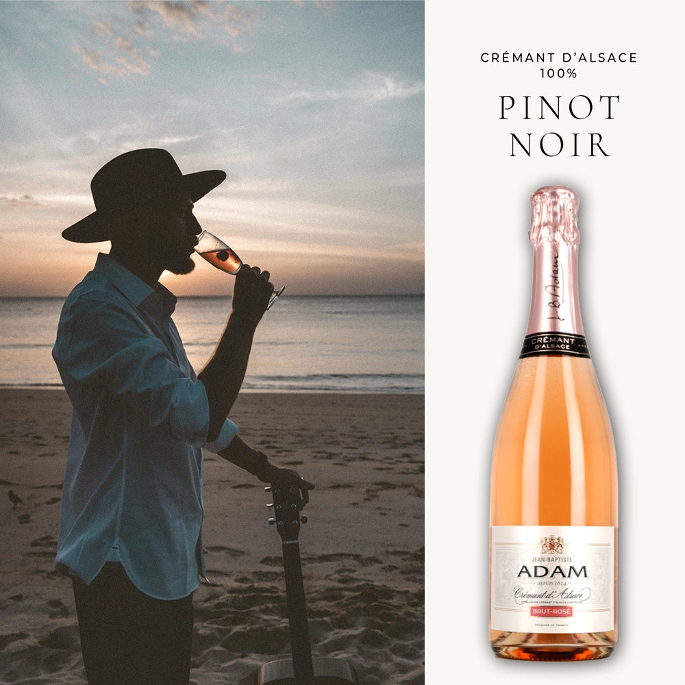 Eine Person am Strand bei Sonnenuntergang, die ein Glas Crémant d'Alsace Brut Rosé genießt. Im Vordergrund steht eine akzentuierte Flasche dieses Schaumweins mit der Aufschrift "100% Pinot Noir" der Marke Jean Baptiste Adam. Das Ambiente vermittelt eine entspannte und genussvolle Stimmung.