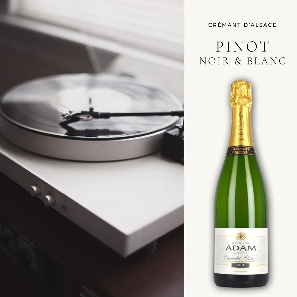 Eine Flasche Crémant d'Alsace, beschriftet mit "Pinot Noir & Blanc" von Jean-Baptiste Adam, vor einem unscharfen Hintergrund mit einem Plattenspieler, auf dem eine Vinylplatte abgespielt wird. Die Flasche steht rechts im Bild und ist scharf im Vordergrund zu sehen, während der Plattenspieler links im Bild durch eine geringe Tiefenschärfe weichgezeichnet ist.