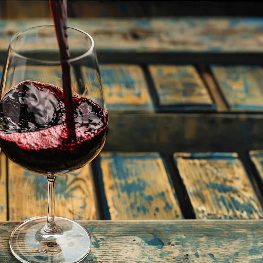 Ein Glas mit Rotwein, der vor einem Hintergrund mit Holztextur steht. In das Glas wird gerade Rotwein eingegossen, wodurch eine dynamische Bewegung im Wein zu sehen ist, die durch die Schlieren und Blasen betont wird. Die Farbe des Weins ist tiefrot, was typisch für einen Côtes du Rhône von Brunel de la Gardine ist.