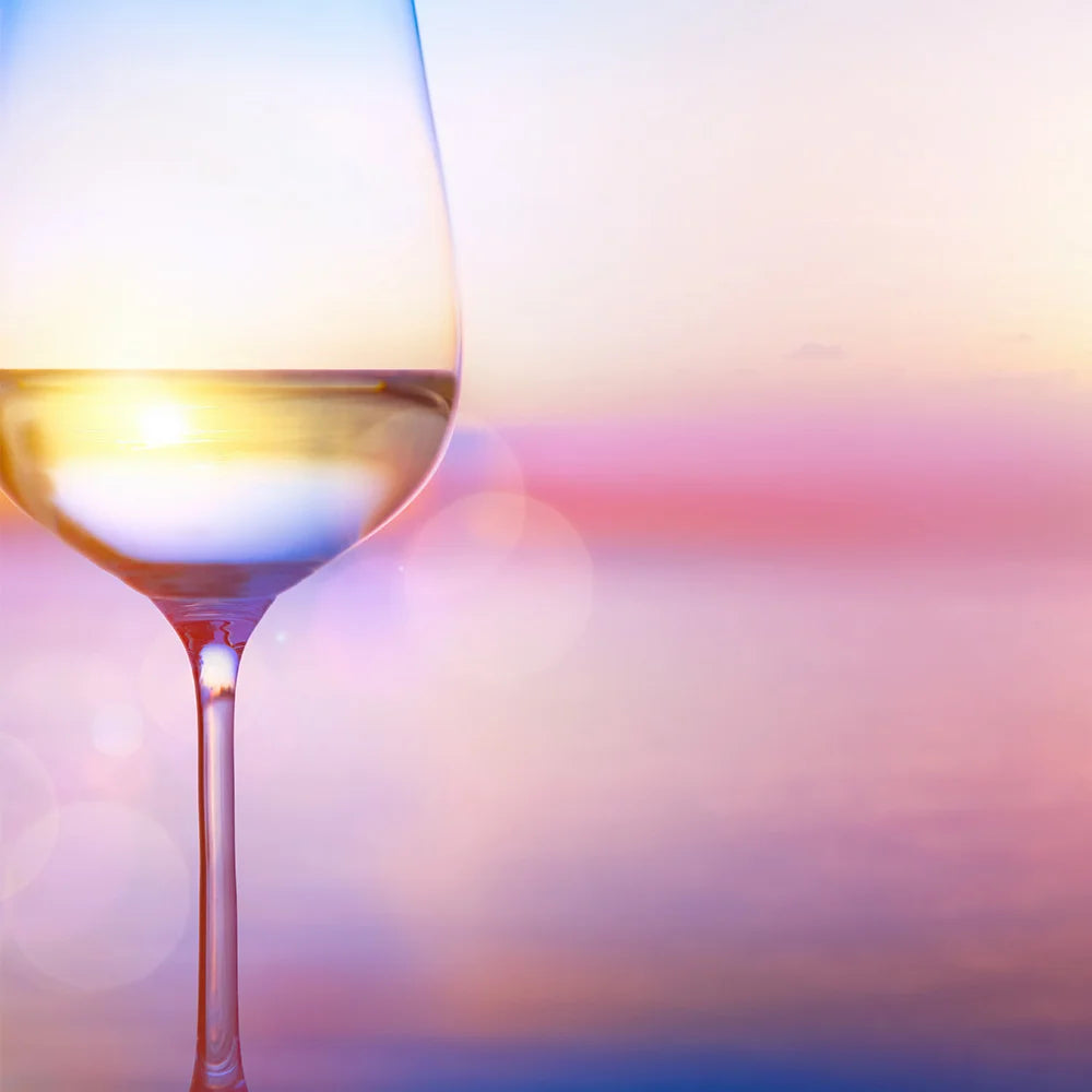 Ein Glas Weißwein, eingefangen vor der Kulisse eines sommerlichen Sonnenuntergangs. Die sanften Pastelltöne des Abendhimmels ergänzen die goldene Klarheit des Weines. 