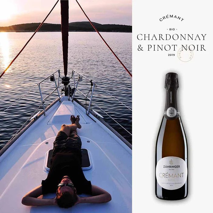 Crémant-Flasche neben einer entspannenden Bootsszene während eines malerischen Sonnenuntergangs auf dem See, symbolisierend den perfekten Genussmoment.