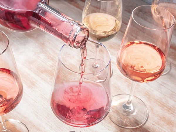 Roséwein wird aus einer Flasche in eines von mehreren Gläsern auf einem Holztisch gegossen, wobei jedes Glas eine unterschiedliche Schattierung des Weines aufweist.